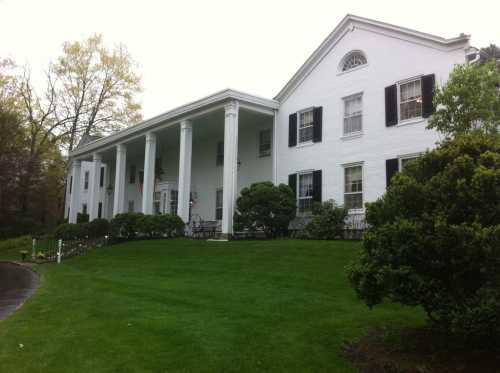 A large white house with columns, black shutters, and a well-manicured lawn surrounded by greenery.