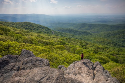 A person stands on a rocky outcrop, overlooking a lush green valley and distant mountains under a bright sky.