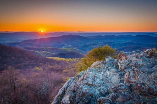 Sunrise over rolling mountains, with vibrant colors illuminating the sky and rocky foreground.