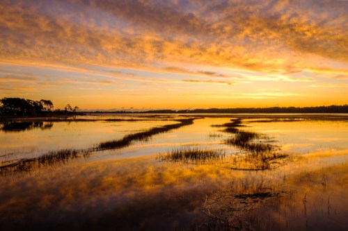 A serene sunset over a calm marsh, with vibrant orange and purple clouds reflecting in the water.