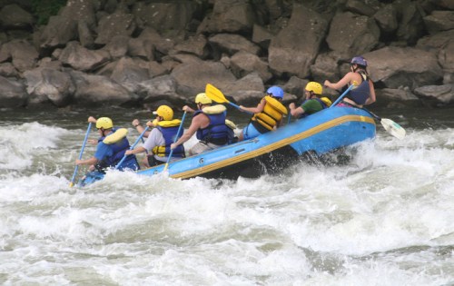 A group of people in a blue raft navigating whitewater rapids, wearing helmets and life jackets.