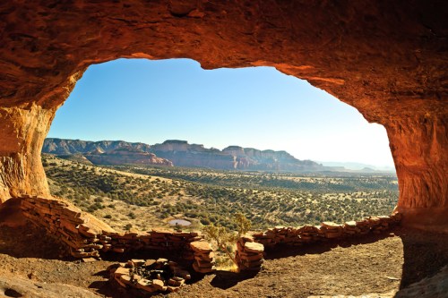 A panoramic view from a cave opening, showcasing a vast desert landscape and distant mountains under a clear blue sky.