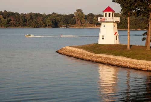 A white lighthouse with a red roof stands by a calm lake, with a boat speeding past and trees in the background.