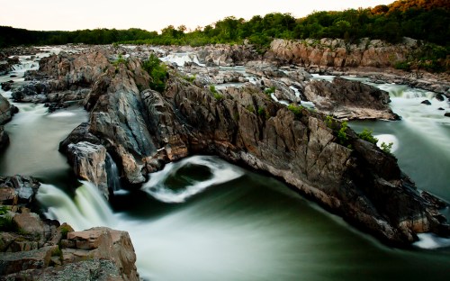 A scenic view of a river flowing over rocky formations, surrounded by lush greenery and a serene landscape.