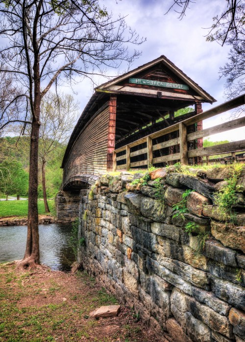 A historic covered bridge stands over a stone wall by a calm river, surrounded by trees and greenery.