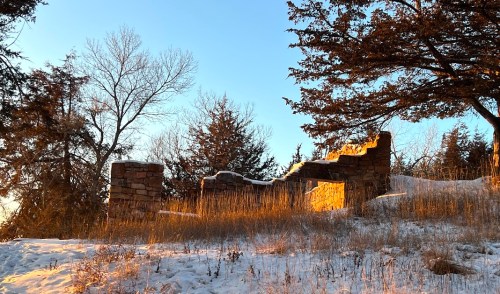 Ruins of a stone structure on a snowy hillside, surrounded by trees, illuminated by warm sunset light.