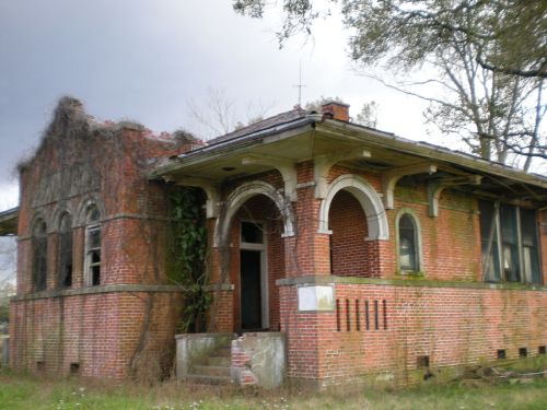 Abandoned brick house overgrown with vines, featuring arched windows and a deteriorating roof, set in a grassy area.