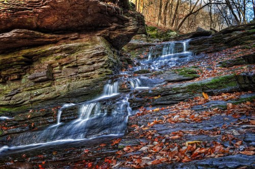 A serene waterfall cascades over rocky terrain, surrounded by autumn leaves and trees in a peaceful forest setting.
