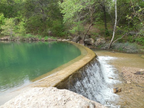 A serene scene of a small waterfall cascading over a stone ledge into a clear, greenish-blue pond surrounded by trees.