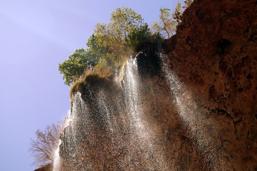 A waterfall cascading down a rocky cliff, surrounded by greenery under a clear blue sky.