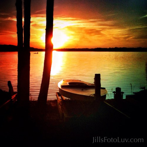 Sunset over a calm lake, with silhouettes of trees and a boat on the shore, creating a serene atmosphere.