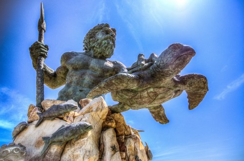 A large statue of a muscular figure holding a spear, riding a turtle, surrounded by fish and set against a bright sky.