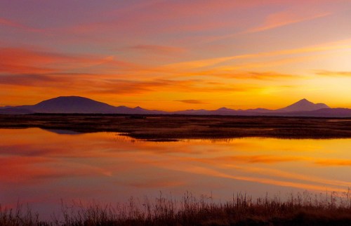 A serene sunset over a calm lake, reflecting vibrant orange and purple hues with distant mountains in the background.