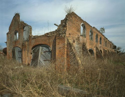 Abandoned brick building with crumbling walls, broken windows, and overgrown grass under a cloudy sky.