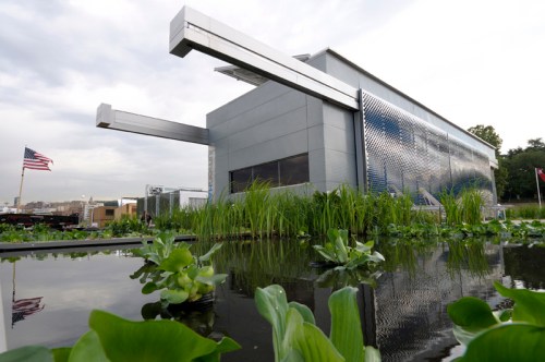 Modern building with a metallic facade, surrounded by greenery and a reflective pond, under a cloudy sky.