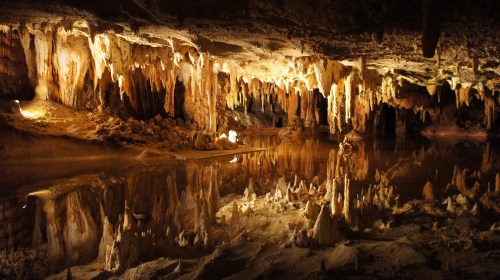 A dimly lit cave with stalactites and stalagmites reflected in a calm underground pool of water.