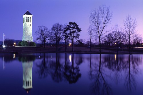 A stone tower beside a calm lake, reflecting trees and a twilight sky in shades of purple and blue.