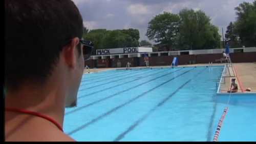 A lifeguard watches over a large, empty swimming pool on a sunny day, with trees in the background.