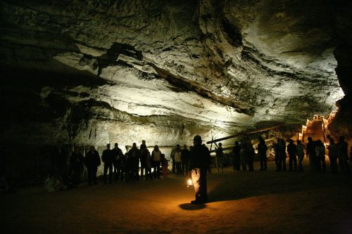 A group of people stands in a dimly lit cave, with a guide holding a lantern in the foreground and stairs in the background.