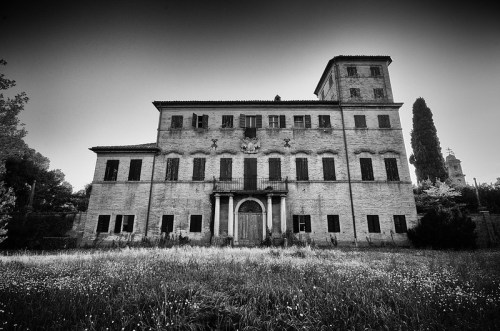 A dilapidated, historic building with a tower, surrounded by overgrown grass and trees, in black and white.
