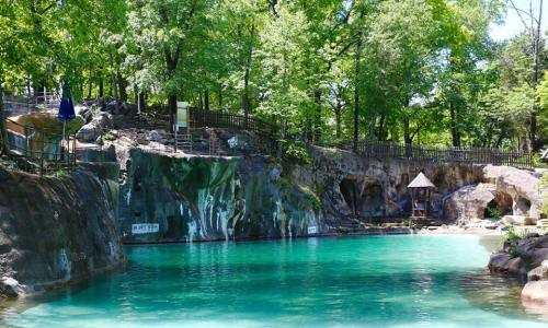 A serene swimming area surrounded by lush greenery and rocky cliffs, with clear turquoise water and a wooden gazebo.
