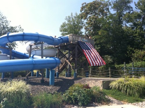 A water slide with a wooden structure, featuring an American flag draped down the side, surrounded by greenery.