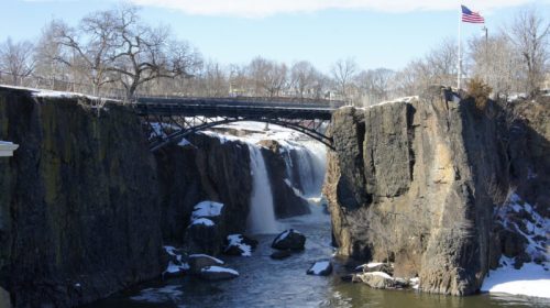 A snowy landscape featuring a waterfall, rocky cliffs, and a bridge with an American flag in the background.