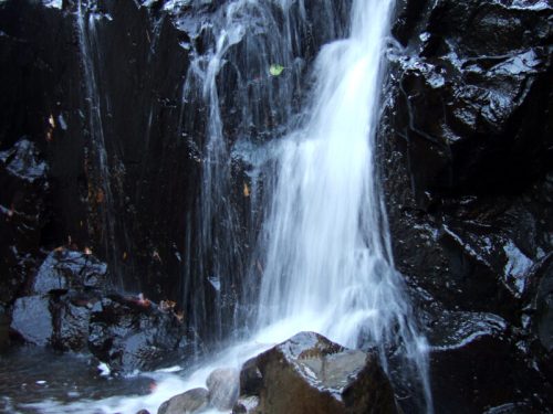 A cascading waterfall flows over dark rocks, surrounded by a serene natural setting.