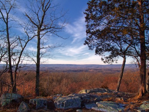 A scenic view from a rocky overlook, featuring trees and a vast landscape under a blue sky with wispy clouds.