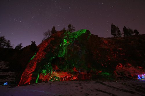 A rocky cave illuminated in red and green lights under a starry night sky. Snow covers the ground.
