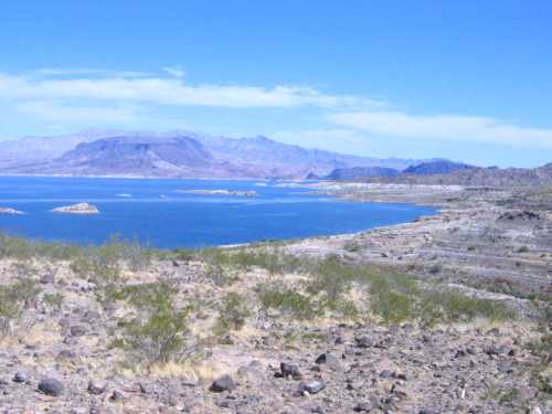 A scenic view of a blue lake surrounded by rocky terrain and distant mountains under a clear blue sky.