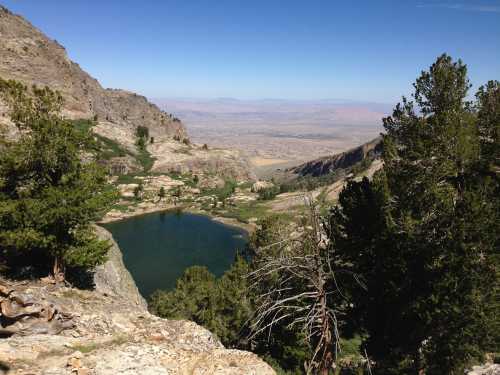 A serene mountain landscape featuring a clear lake surrounded by rocky terrain and tall trees under a blue sky.