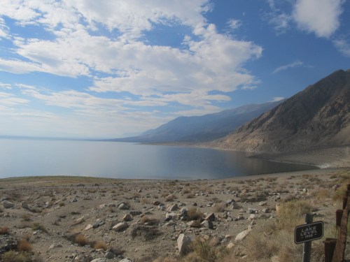 A serene lake surrounded by mountains under a partly cloudy sky, with rocky terrain in the foreground.