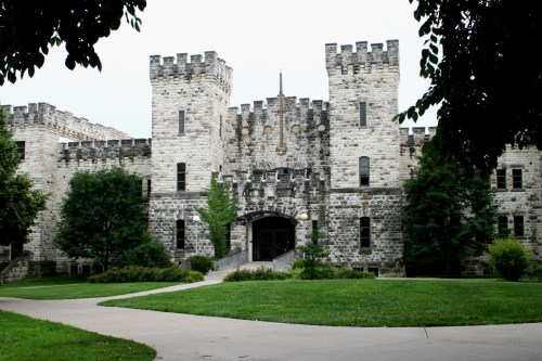 A stone castle with towers and a central entrance, surrounded by green grass and trees.