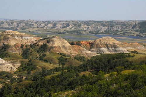 A panoramic view of rolling hills and layered rock formations under a clear blue sky, with greenery in the foreground.