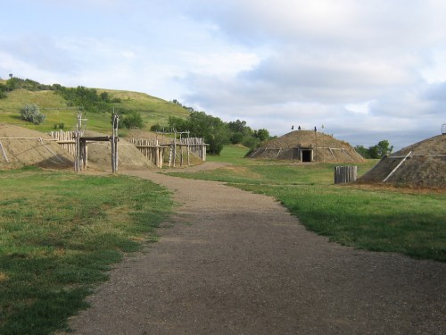 A dirt path leads through grassy terrain with two earthen mounds and wooden structures under a cloudy sky.