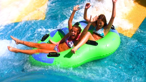 Two girls joyfully sliding down a water slide on a green inflatable tube, splashing in the blue water below.