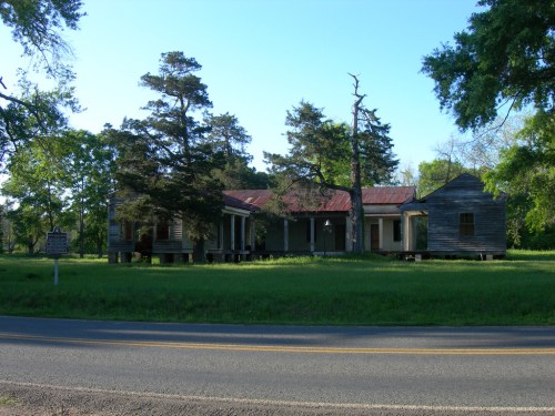Abandoned wooden house with a red roof, surrounded by trees, set against a clear blue sky and green grass.