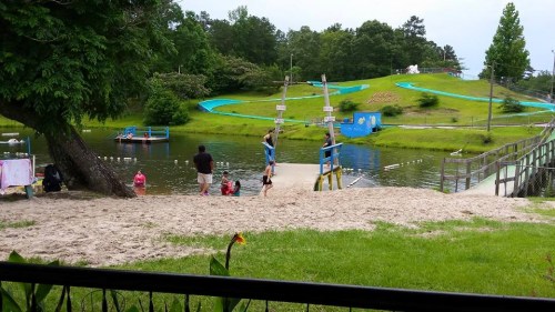 A sandy area by a lake with children playing, water slides in the background, and green hills surrounding the scene.