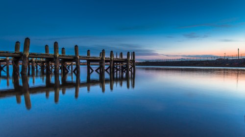 A serene pier extends into calm waters at dusk, reflecting the blue sky and soft sunset hues.