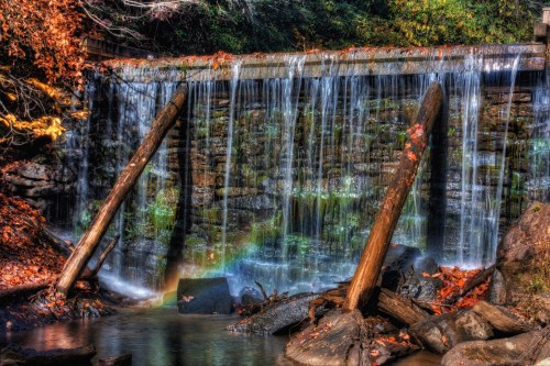 A serene waterfall cascades over a stone wall, surrounded by autumn foliage and wooden logs in a tranquil setting.