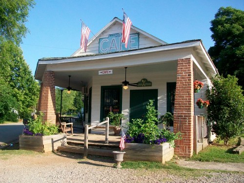 A charming café with American flags, surrounded by flowers and greenery, set in a sunny outdoor scene.