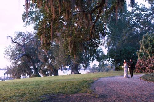 A couple walks hand in hand along a path under large trees, with a serene lake in the background at dusk.