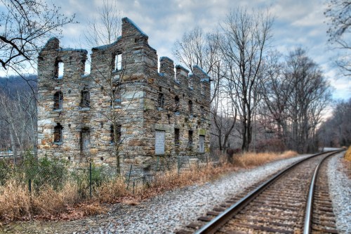 A weathered stone ruin beside a gravel path and railway tracks, surrounded by bare trees and a cloudy sky.