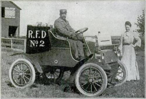 A man sits in an early motor vehicle labeled "R.F.D. No. 2," while a woman stands beside him in a rural setting.