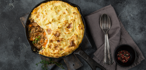 A cast iron skillet with shepherd's pie, featuring a golden mashed potato topping and a portion removed, on a dark surface.