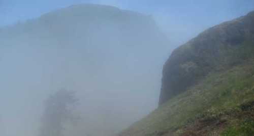 A misty landscape with a rocky hill partially obscured by fog and a hint of greenery in the foreground.