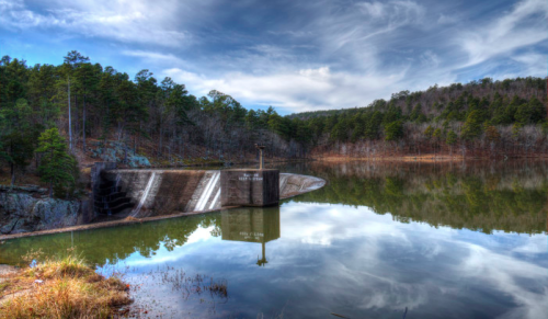 A serene landscape featuring a dam reflecting in calm water, surrounded by trees and a cloudy sky.
