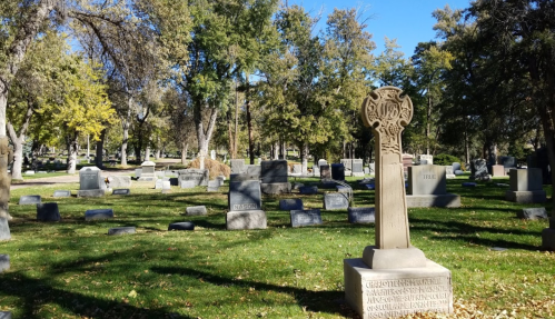 A serene cemetery scene with a decorative gravestone surrounded by trees and other headstones under a clear blue sky.