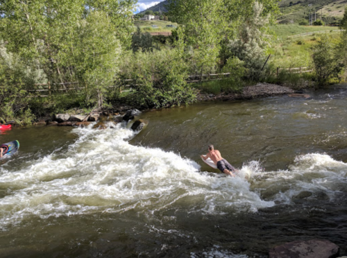 A person is surfing on a river wave, surrounded by greenery and rocky banks. Another kayaker is visible nearby.